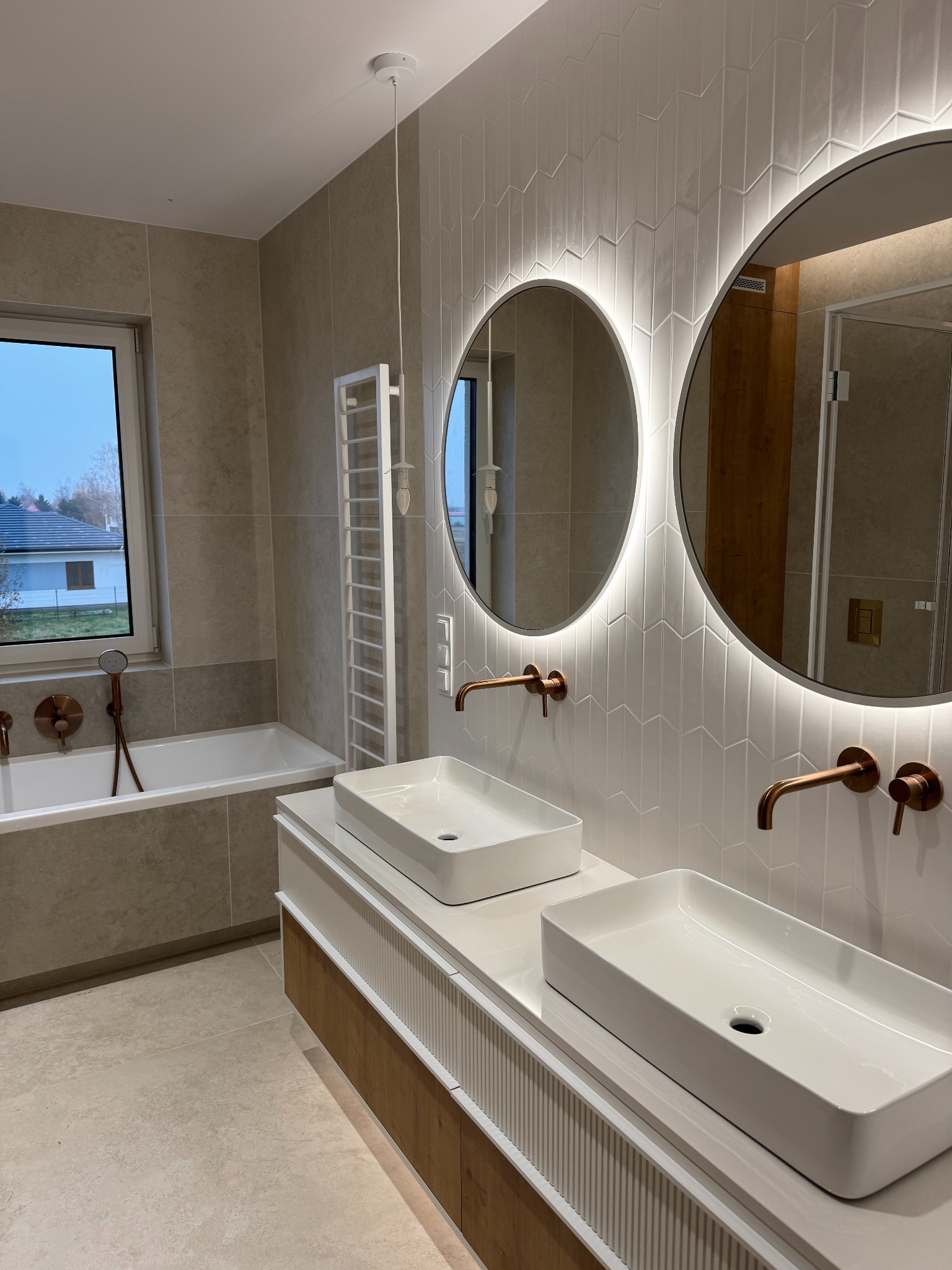 Nowoczesna łazienka z dwoma umywalkami nablatowymi, okrągłymi lustrami z podświetleniem LED i armaturą w kolorze miedzi. Uwagę zwracają białe płytki ułożone w jodełkę.