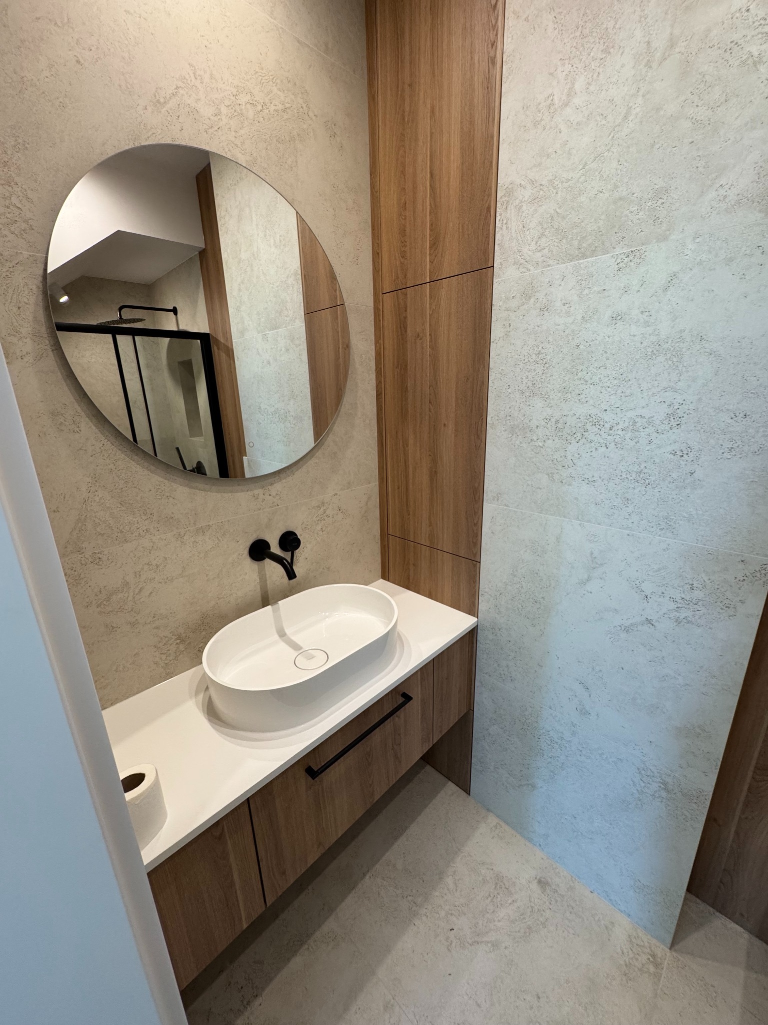 Nowoczesna łazienka z owalną umywalką nablatową, czarną armaturą i okrągłym lustrem. Widoczna kabina prysznicowa w tle. Neutralna kolorystyka płytek i drewnopodobne wykończenie.