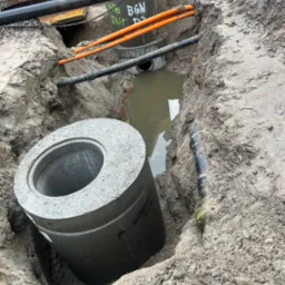 Betonowy krąg w wykopie, obok rury i woda. Widoczne oznaczenia na rurach. Prace ziemne związane z instalacją podziemną.