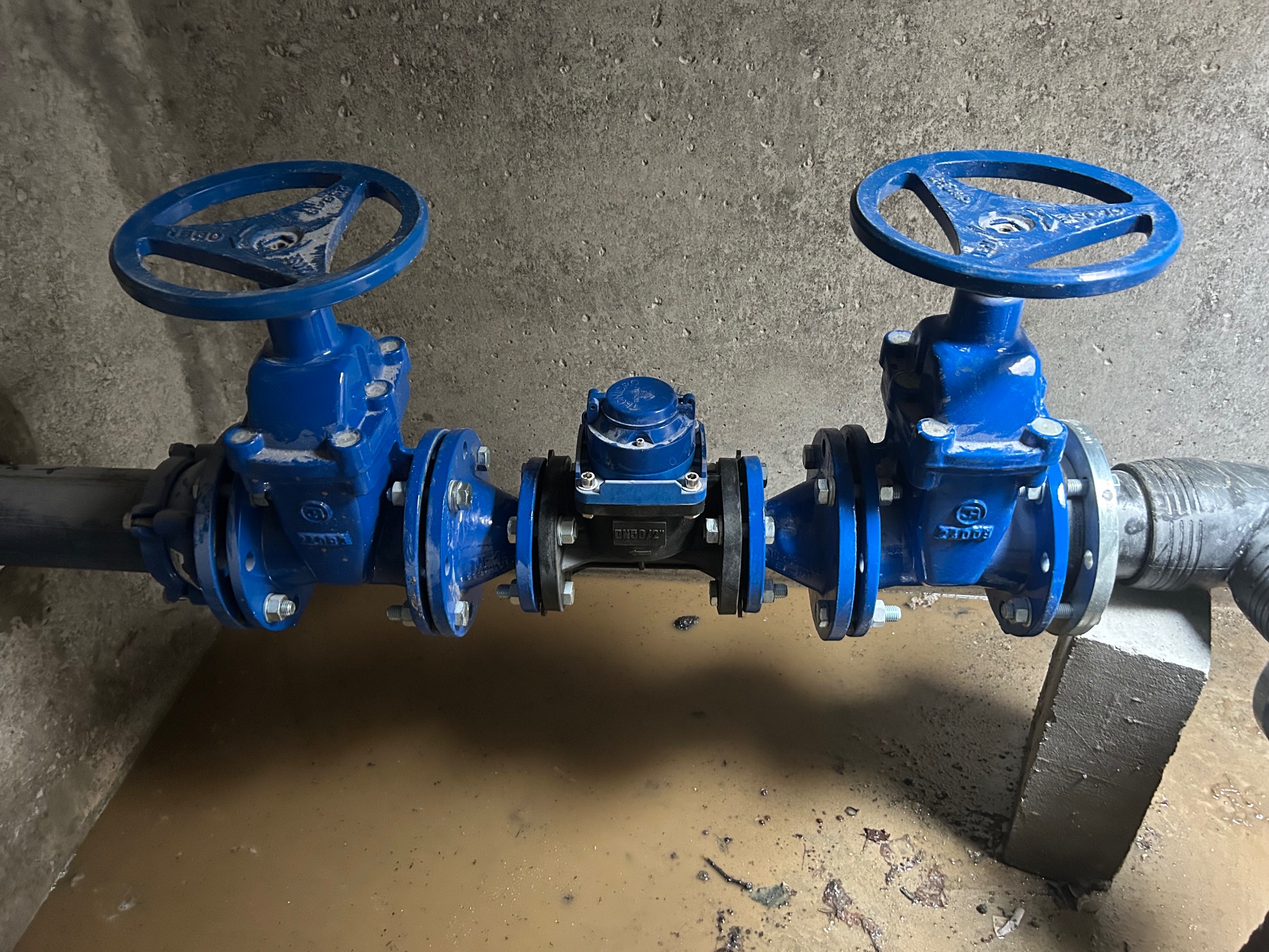 Niebieskie zawory i czarny wodomierz połączone rurami w pomieszczeniu z betonową ścianą. Woda na podłodze. Instalacja wodno-kanalizacyjna.