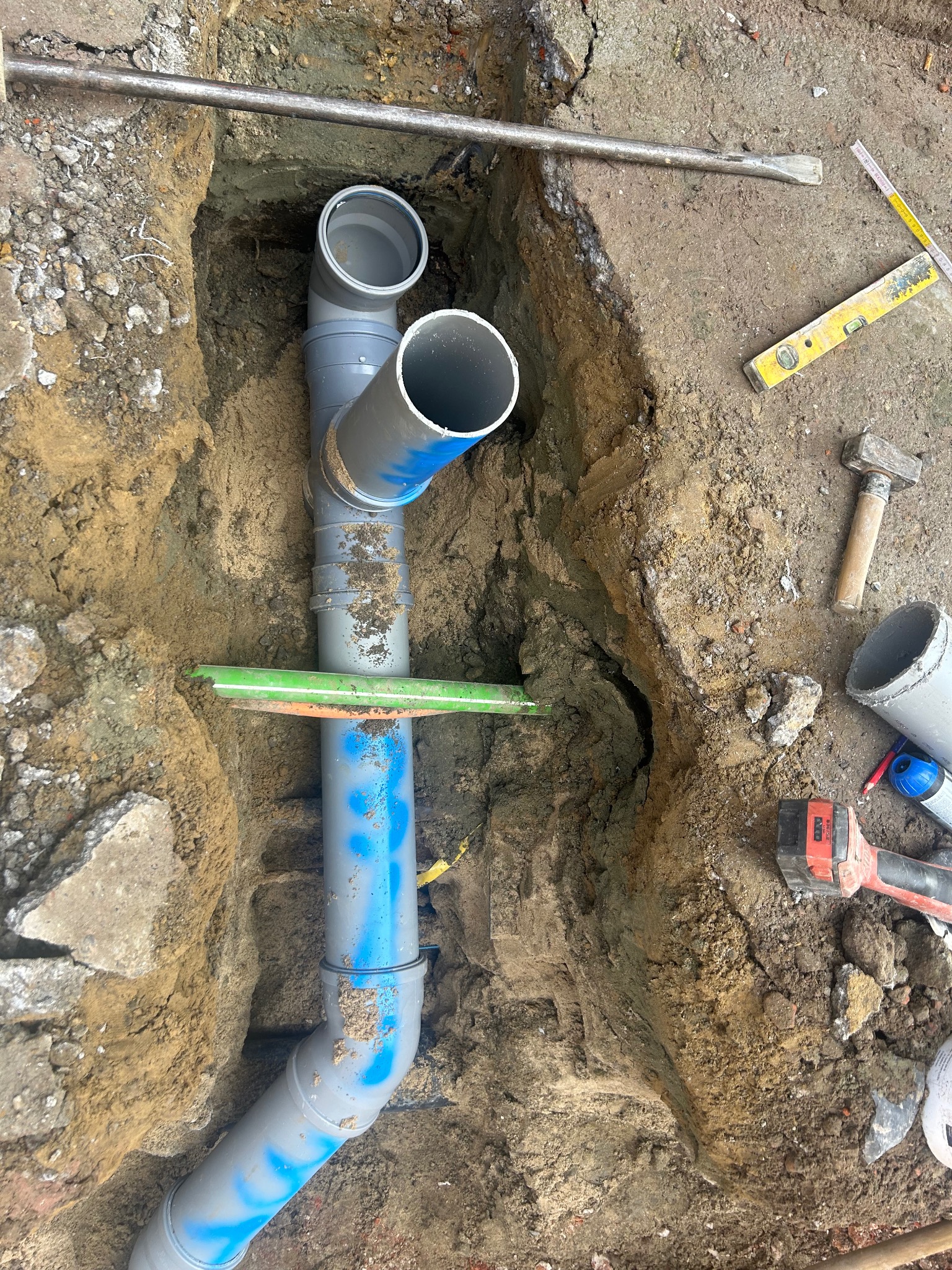 Szare rury kanalizacyjne w wykopie ziemnym, widoczne narzędzia: poziomica, młotek, wiertarka. Elementy instalacji oznaczone niebieskim sprayem.