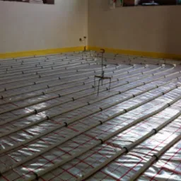 Rozłożone rury ogrzewania podłogowego na folii z czerwoną kratką, przymocowane czarnymi uchwytami, widoczne metalowe stojaki do mocowania rur i żółta taśma przy ścianie.