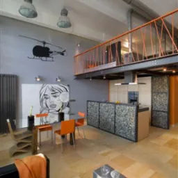 Jasna, kwadratowa posadzka w nowoczesnym, dwupoziomowym wnętrzu z oranżowymi akcentami, widoczne schody na antresolę z pomarańczową balustradą, ściana ozdobiona grafiką i sylwetką helikoptera.