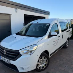 Biały samochód dostawczy Dacia z logo firmy APV na tle nowoczesnego garażu i kostki brukowej. Widoczny numer rejestracyjny PO 6KL83.