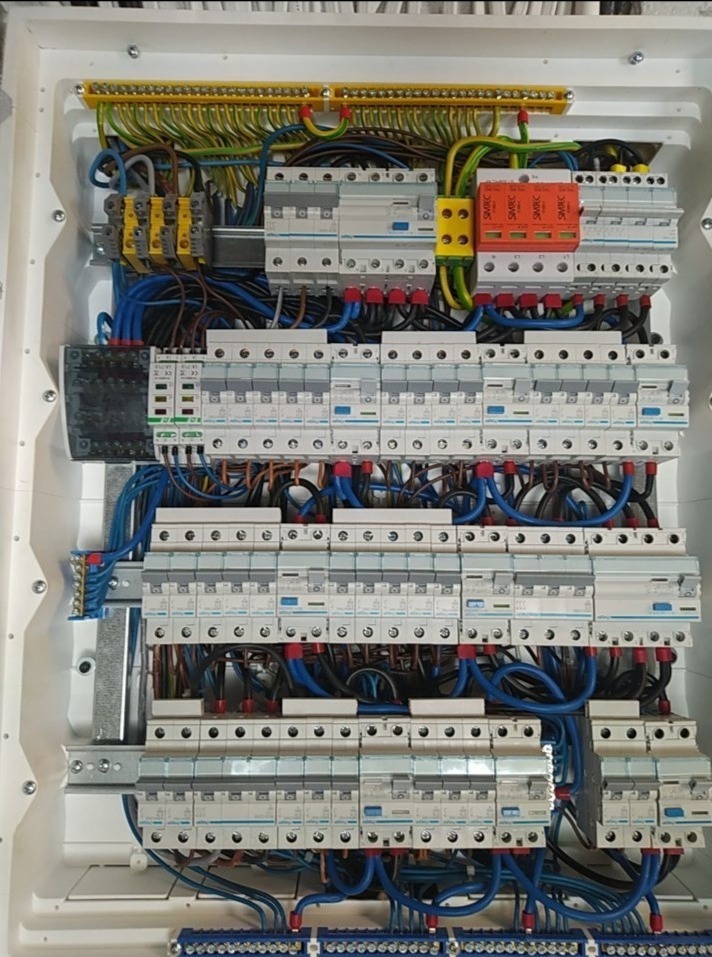 Rozdzielnica elektryczna w białej obudowie, z licznymi bezpiecznikami i przewodami w kolorach niebieskim, czarnym i zielono-żółtym, zamontowana wewnątrz budynku.