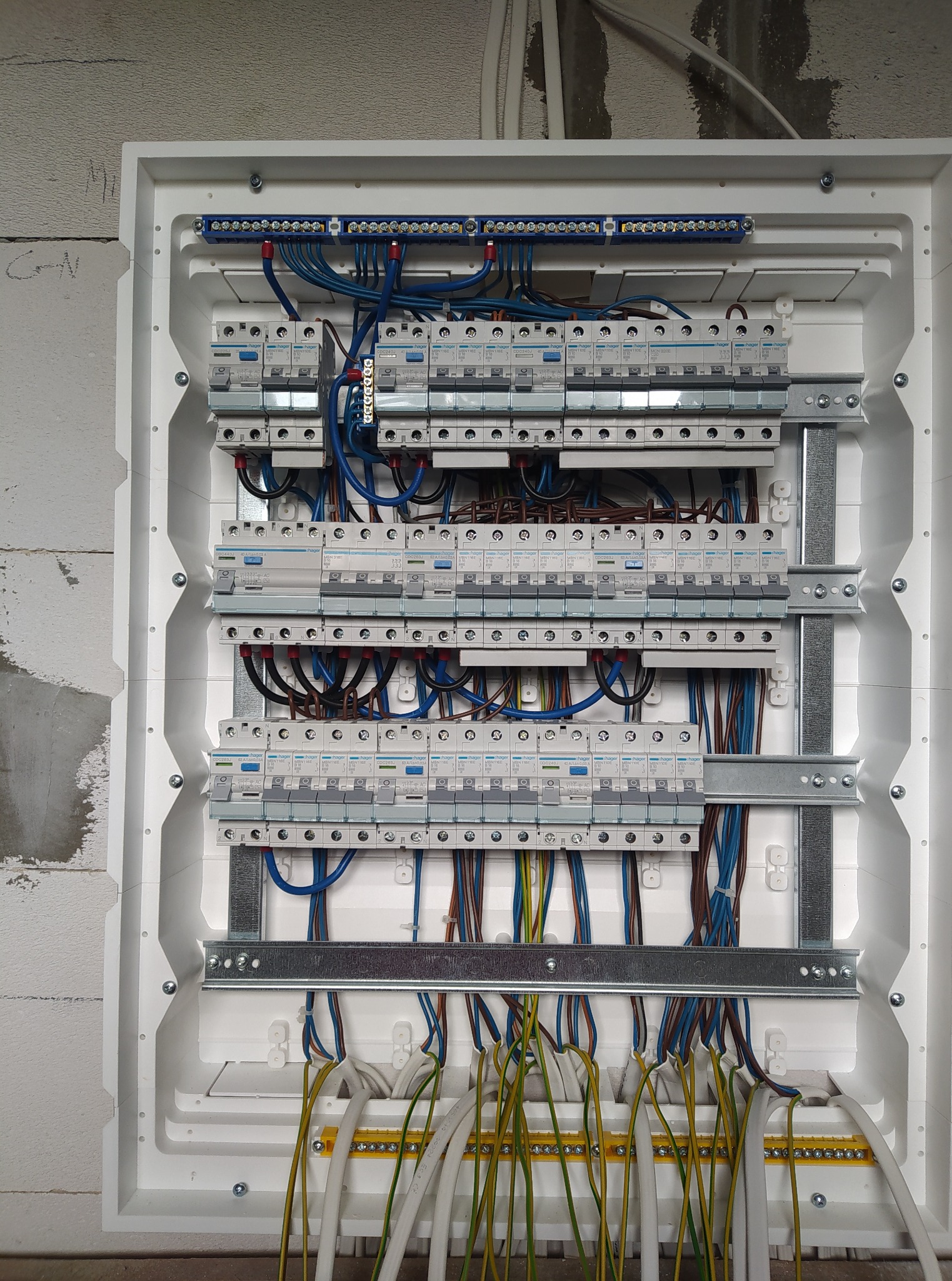 Rozdzielnia elektryczna Hager w białej obudowie, widoczne zabezpieczenia, przewody w kolorach: niebieskim, brązowym, żółto-zielonym. Montaż na ścianie z betonu komórkowego.