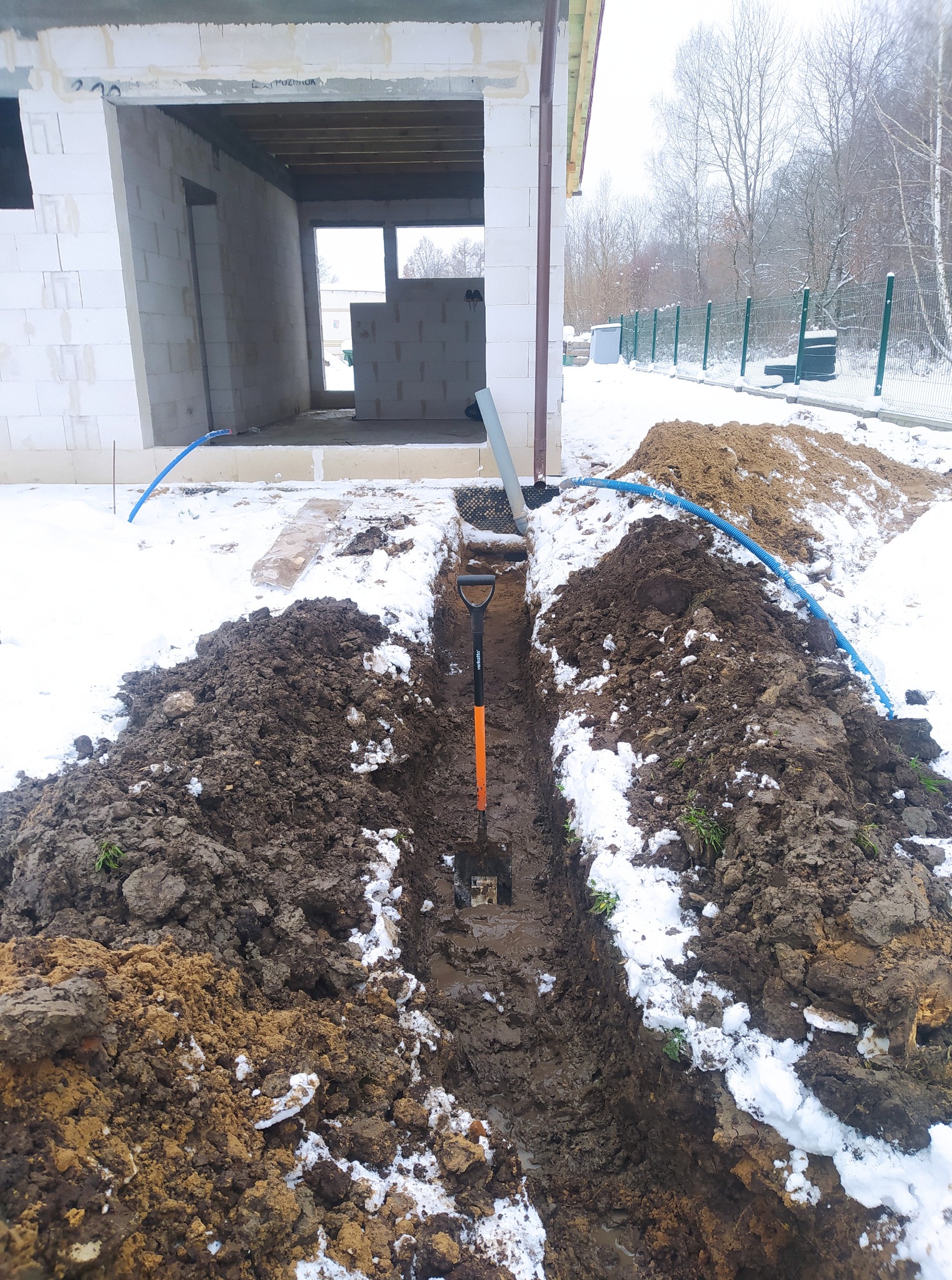Wykop pod kable elektryczne w ziemi, widoczna łopata, niebieski kabel i budynek w trakcie budowy. Zimowy krajobraz z resztkami śniegu.