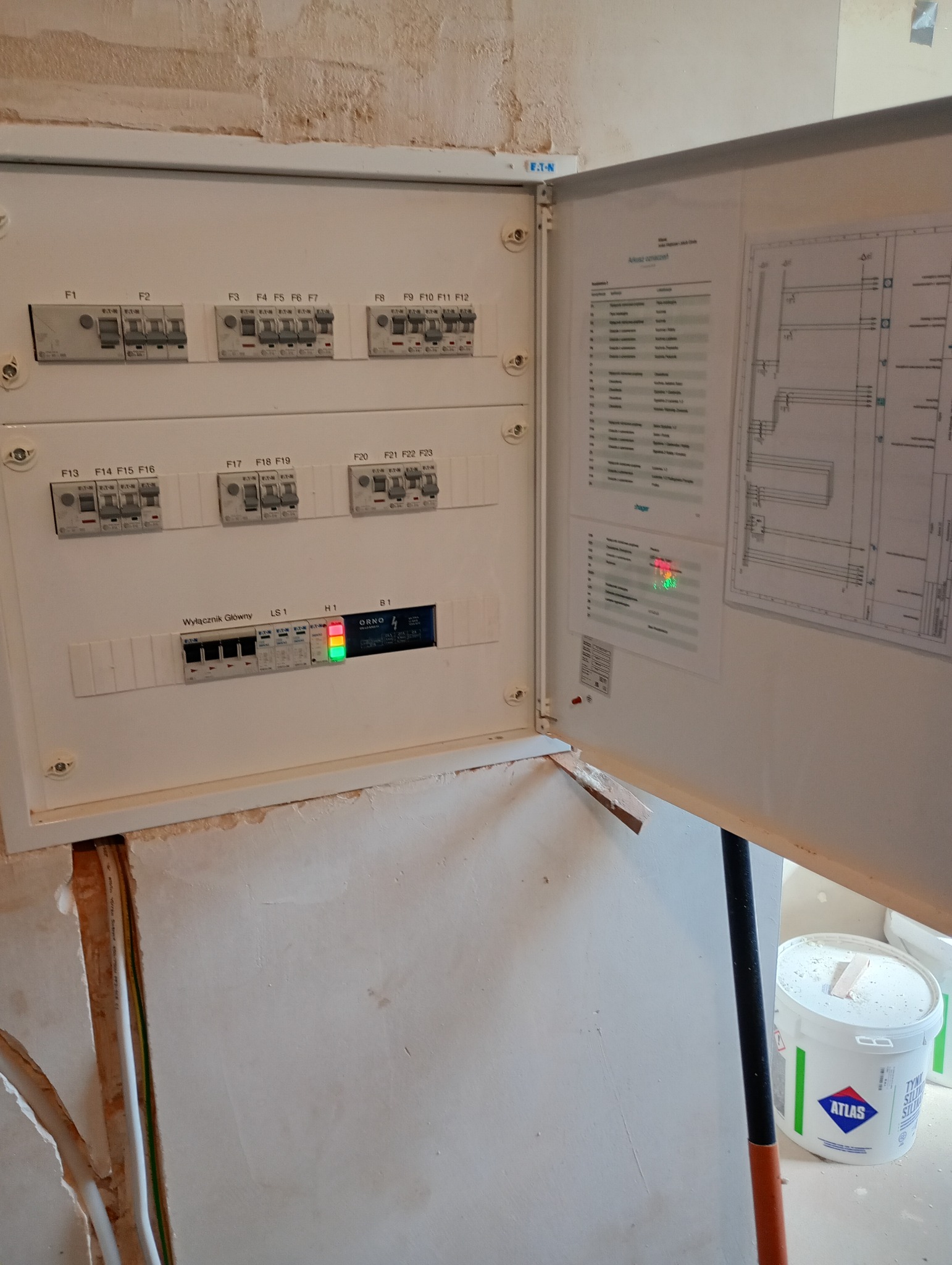 Otwarta skrzynka z bezpiecznikami Eaton i Orno, widoczna instalacja elektryczna, schemat na drzwiczkach. Część ściany w stanie surowym.