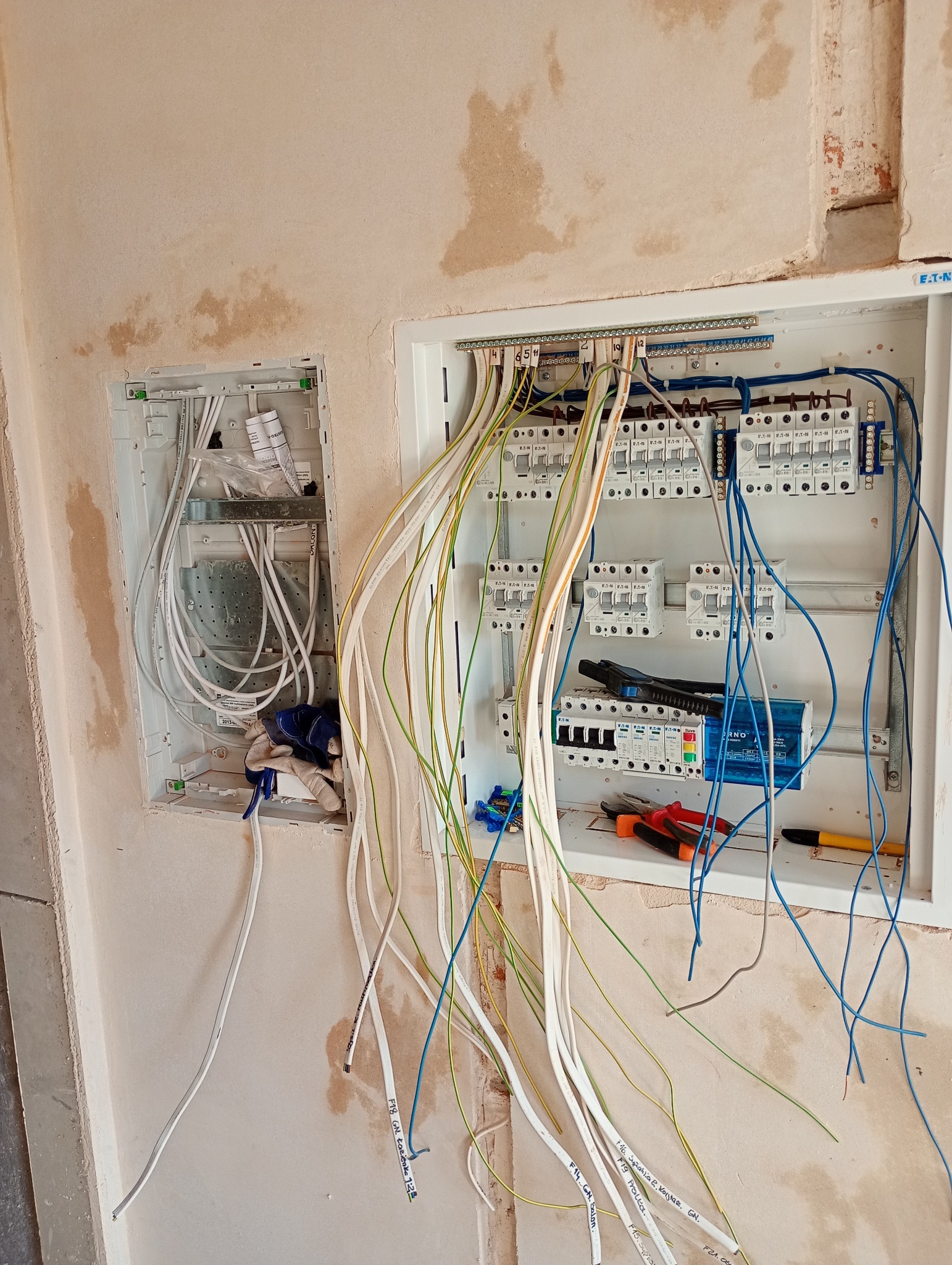 Rozdzielnica elektryczna w trakcie montażu, widoczne kable z oznaczeniami, narzędzia i rękawice robocze. Ściana w stanie surowym.