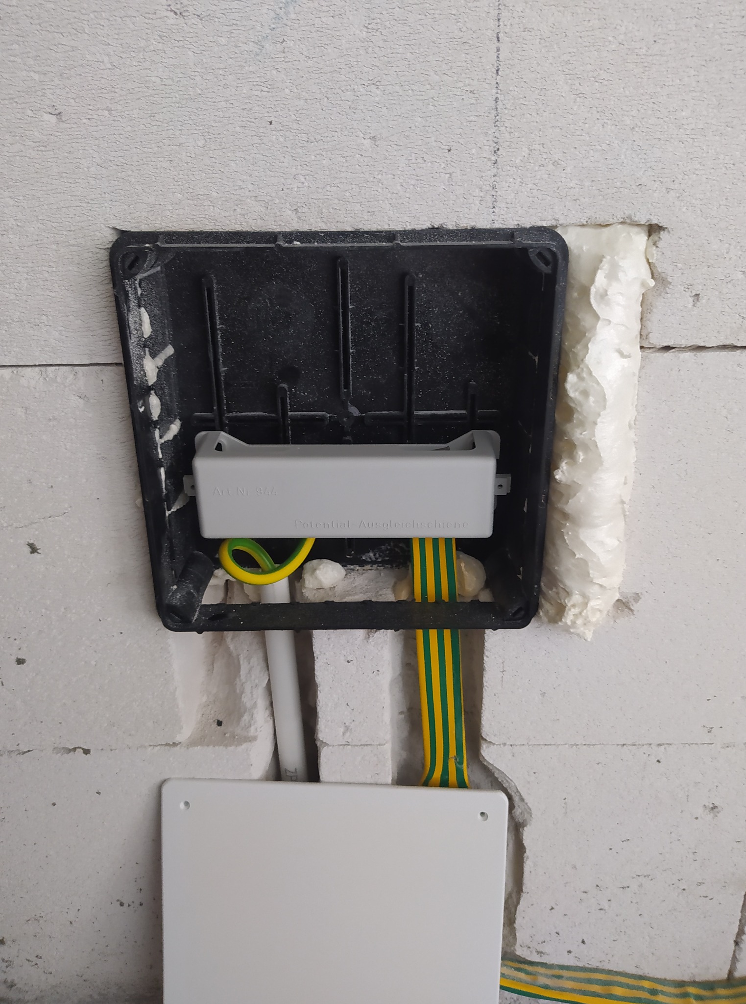 Puszka instalacyjna wmurowana w ścianę z kablami ochronnymi, zabezpieczona pianką montażową. Widoczne elementy instalacji elektrycznej w surowym stanie.