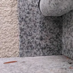 Detale wykończenia elewacji: połączenie szorstkiego tynku w odcieniu beżu i płyt z szarego, nakrapianego granitu. Widoczny narożnik i fragment parapetu.