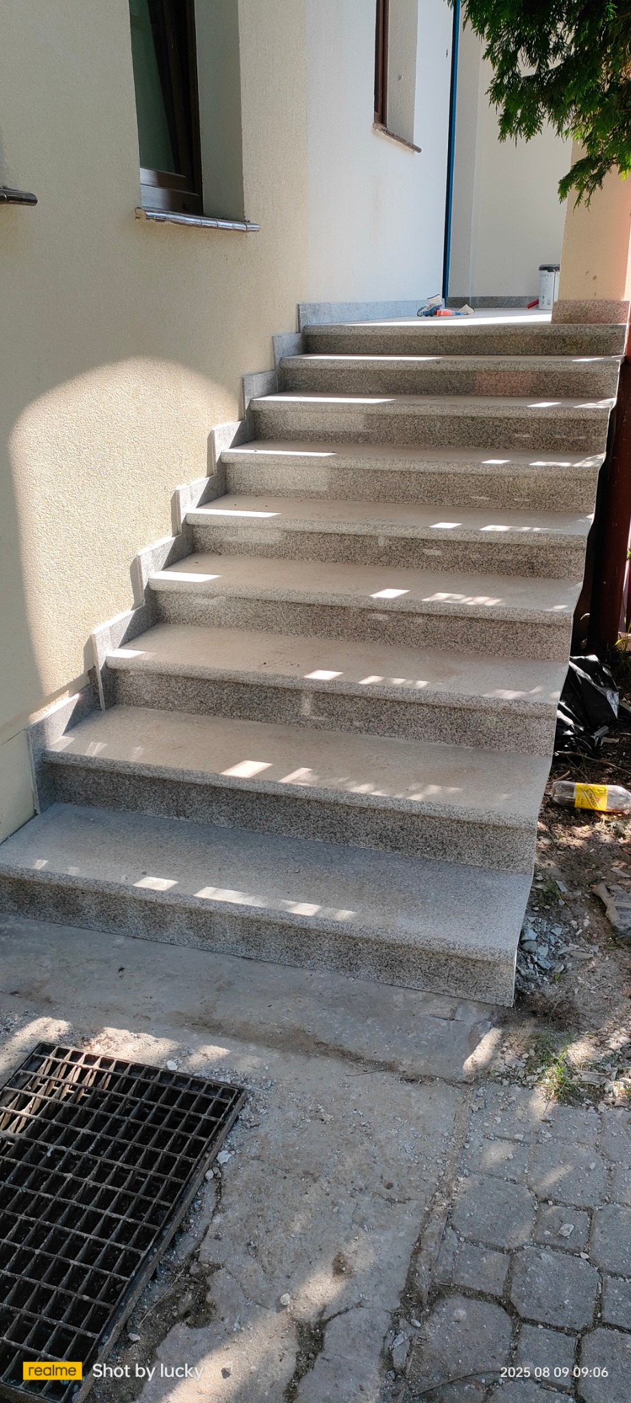 Wykończone schody zewnętrzne z szarego kamienia prowadzące do budynku z beżową elewacją. Widoczny fragment okna i kratka ściekowa.
