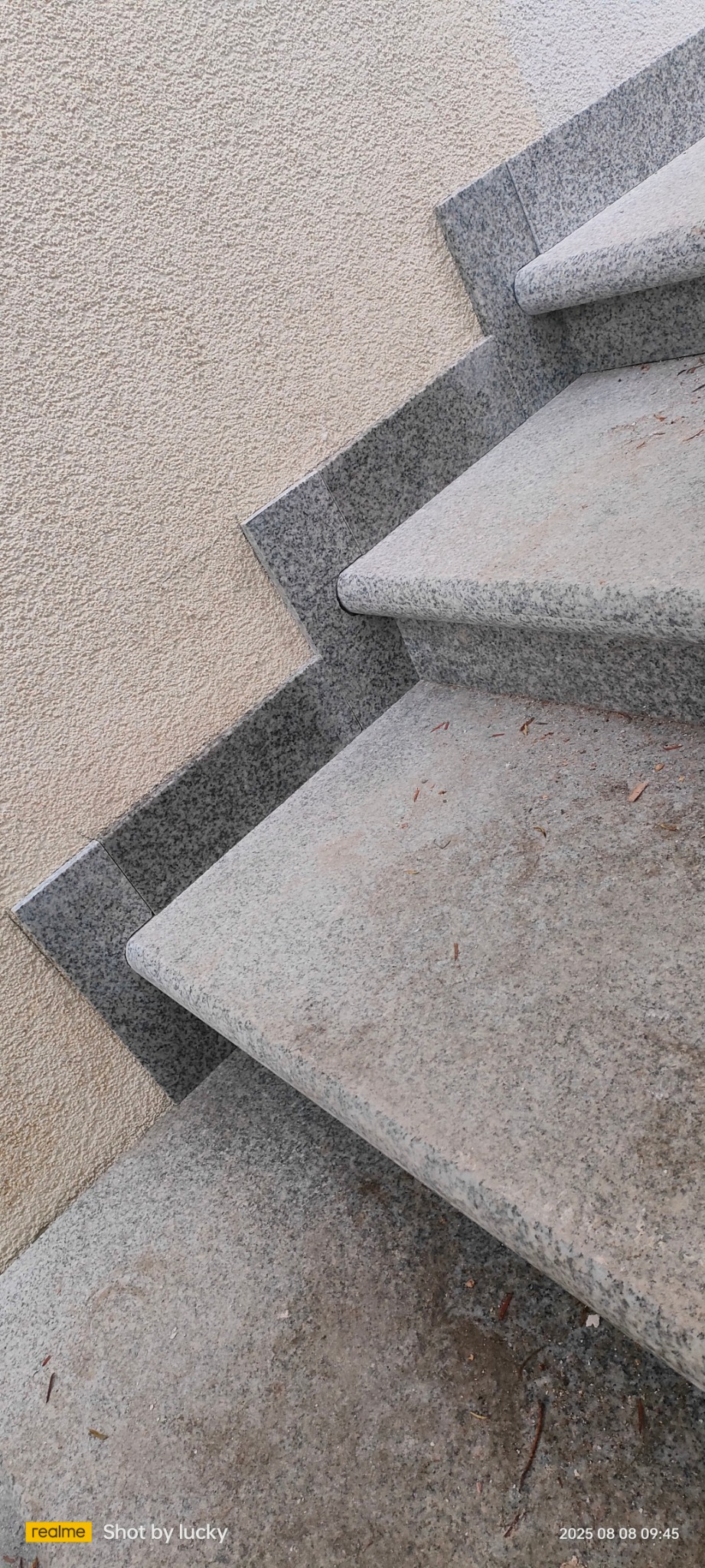 Granitowe schody zewnętrzne z zaokrąglonymi krawędziami, przylegające do ściany o szorstkiej fakturze. Widoczne drobne zanieczyszczenia na stopniach i dokładne wykończenie.