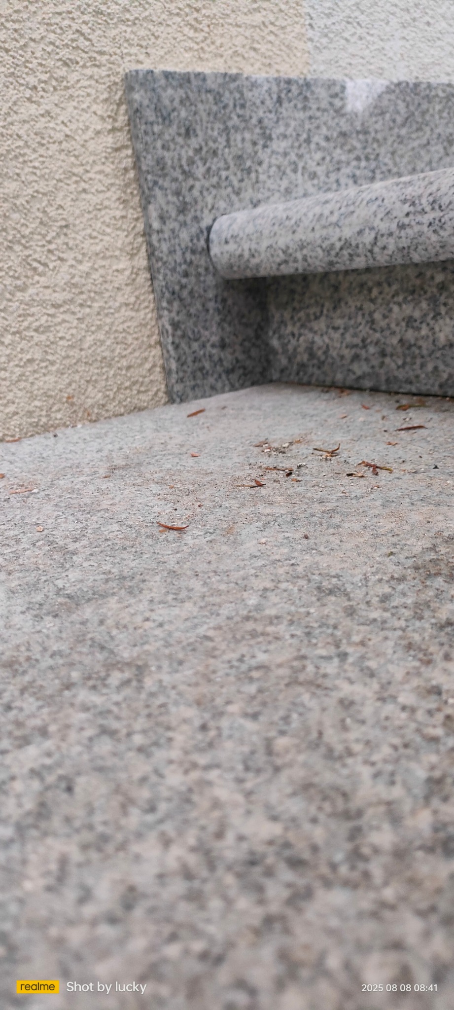 Szpachla murarska z szarego granitu oparta o beżową, chropowatą ścianę. Na szpachlowanej powierzchni leżą drobne, brązowe fragmenty organiczne.