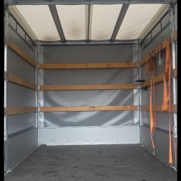 Mat Logis - Wnętrze paki busa transportowego z drewnianymi listwami zabezpieczającymi ładunek i pasami mocującymi, widok z tyłu pojazdu.