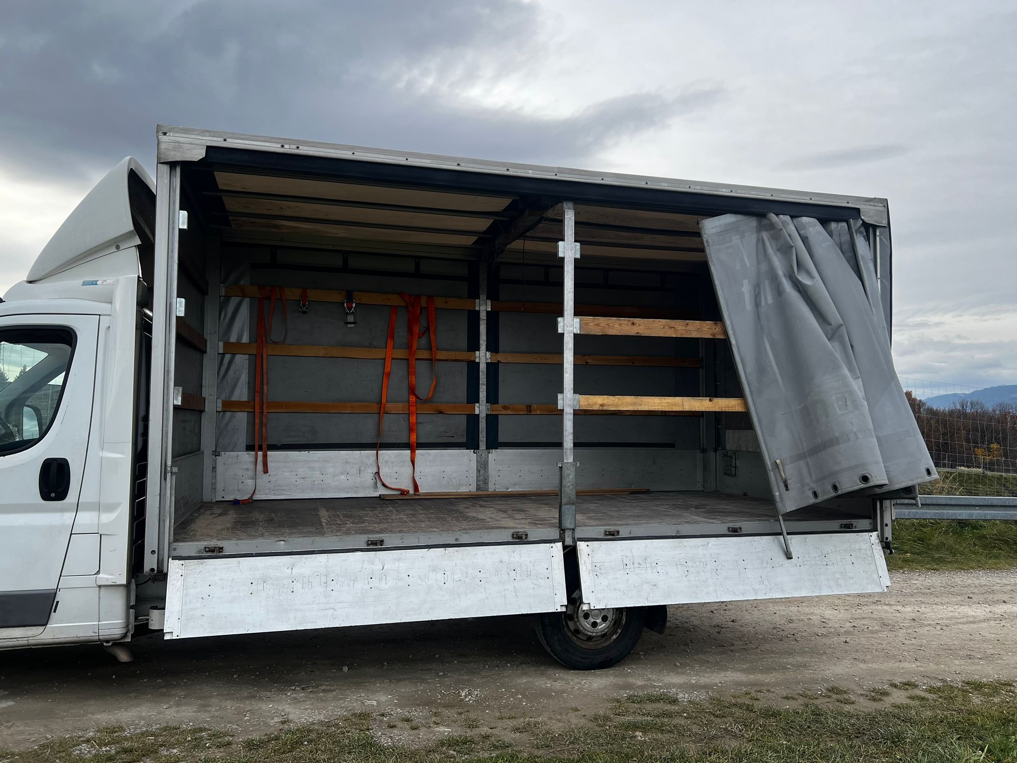 Otwarta paka białego busa z drewnianymi półkami i pomarańczowymi pasami mocującymi, widok z boku na tle pochmurnego nieba i zieleni.