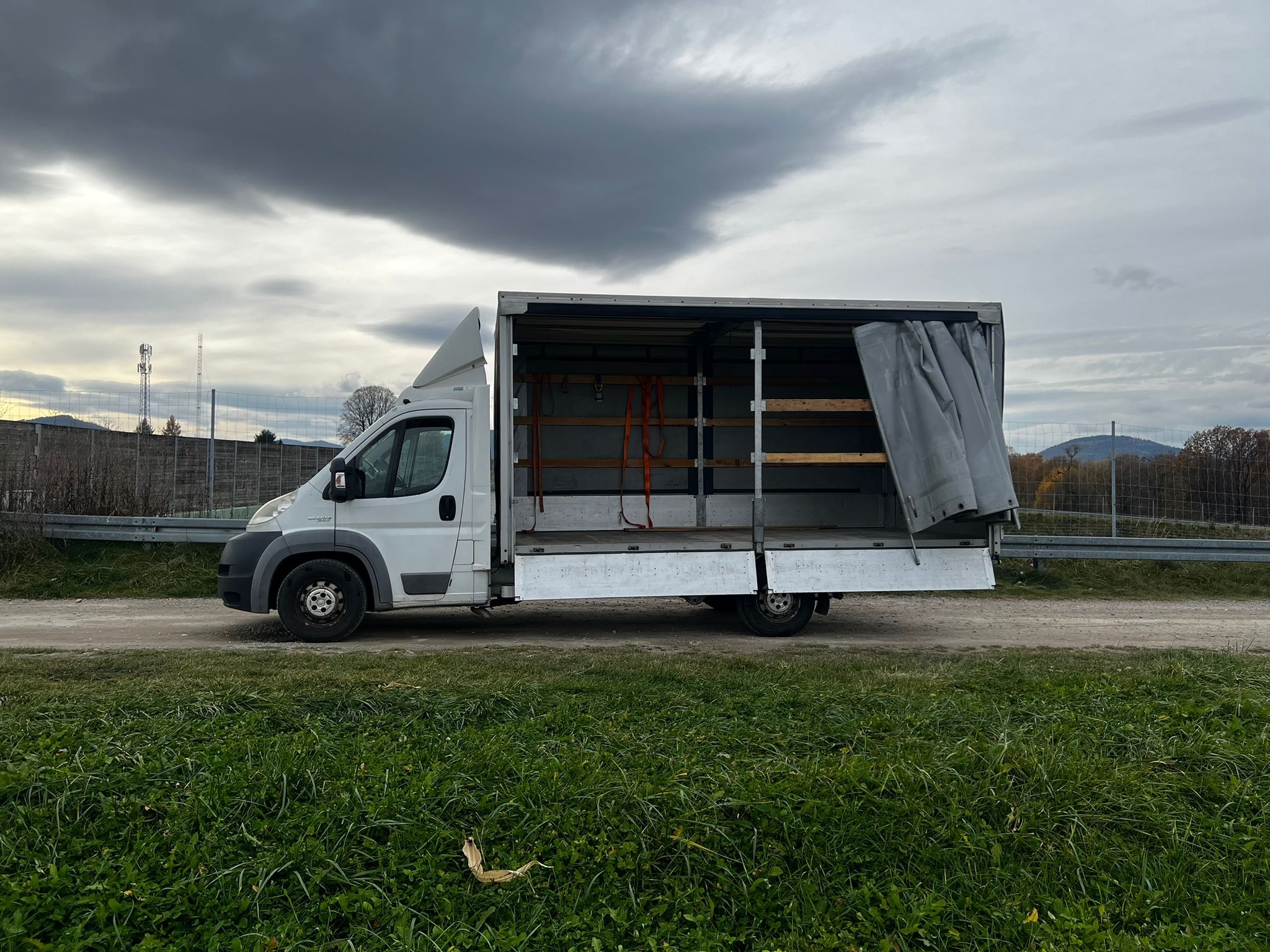 Biały bus z otwartą przestrzenią ładunkową na tle pochmurnego nieba i zielonej trawy. Widoczne pasy mocujące wewnątrz. Krajobraz wiejski w tle.