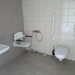 Nowa łazienka dla niepełnosprawnych: białe płytki, składane krzesełko prysznicowe, uchwyty ścienne, toaleta. Funkcjonalne i bezpieczne rozwiązanie.