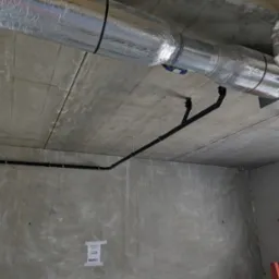 Surowy strop z instalacją: rury wentylacyjne w srebrnej izolacji i czarne rury hydrauliczne. Widoczne mocowania i fragment okna. Szare, betonowe ściany w tle.