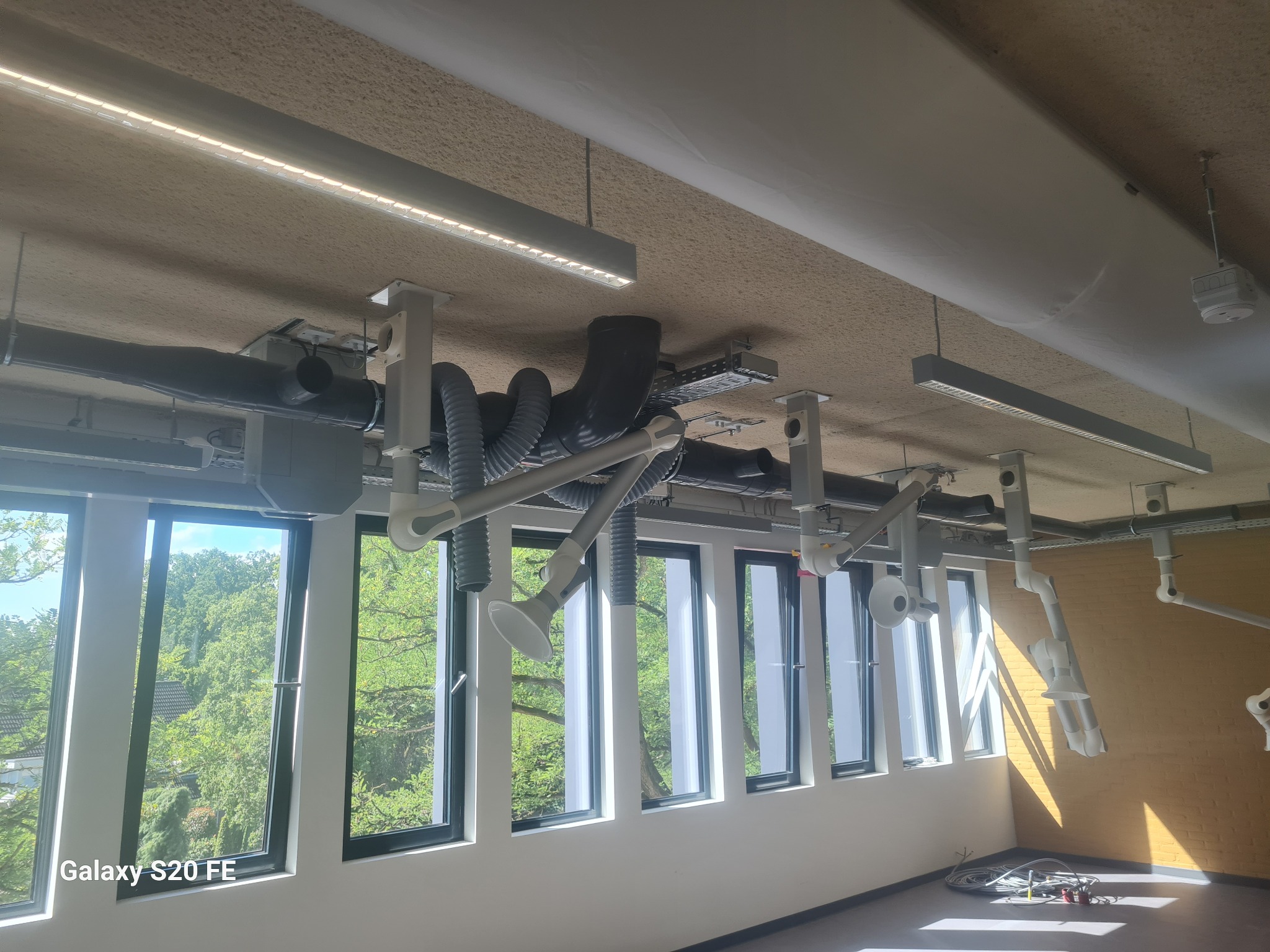 Nowoczesna wentylacja przemysłowa: szare rury i białe ramiona ssące zamontowane pod sufitem w jasnym pomieszczeniu z oknami.