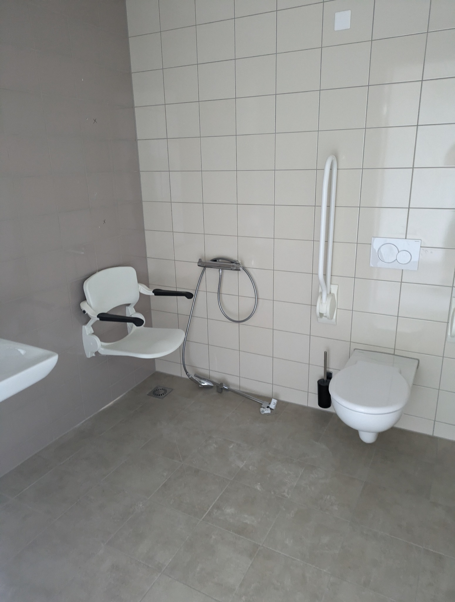Nowa łazienka dla niepełnosprawnych: białe płytki, składane krzesełko prysznicowe, uchwyty ścienne, toaleta. Funkcjonalne i bezpieczne rozwiązanie.