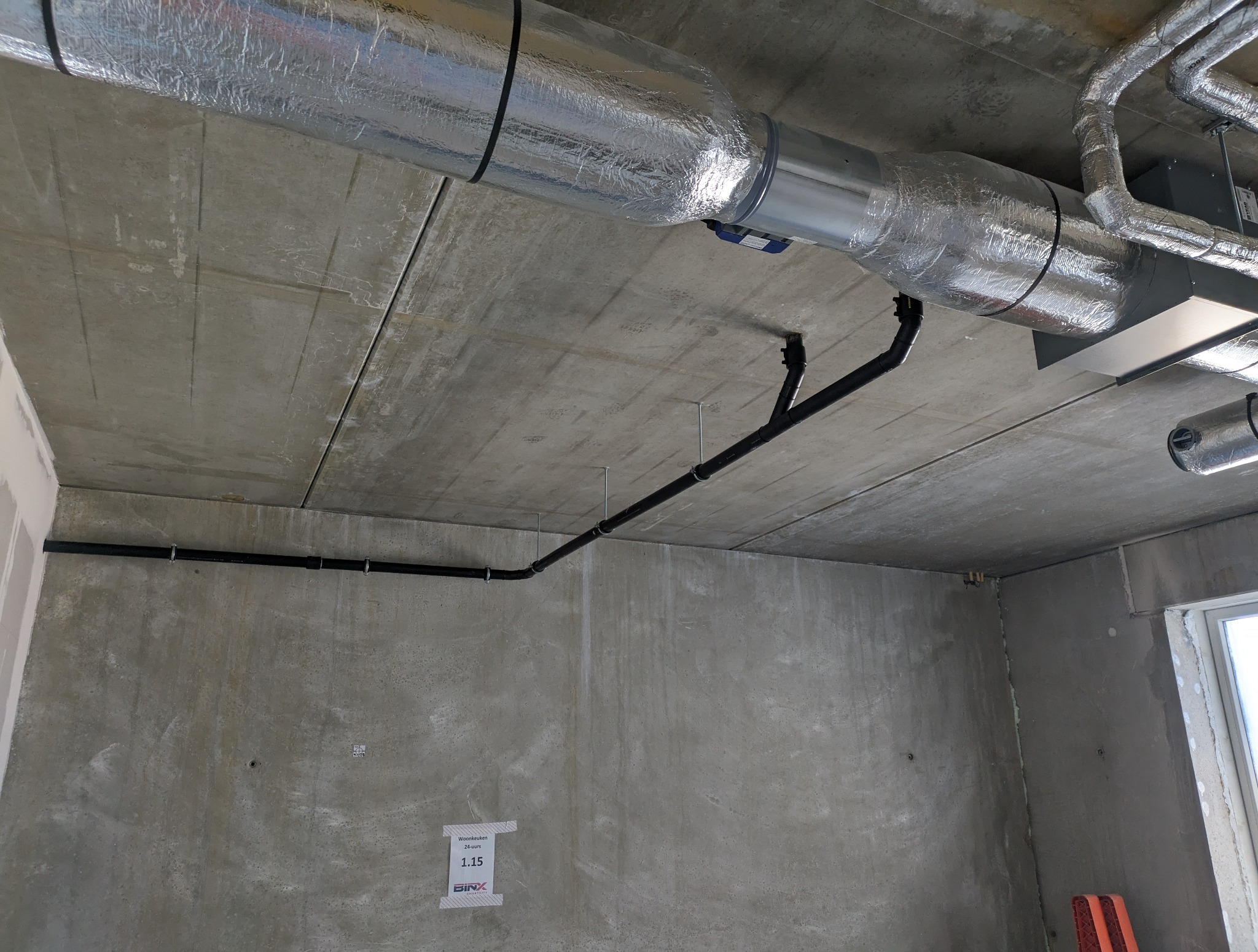 Surowy strop z instalacją: rury wentylacyjne w srebrnej izolacji i czarne rury hydrauliczne. Widoczne mocowania i fragment okna. Szare, betonowe ściany w tle.