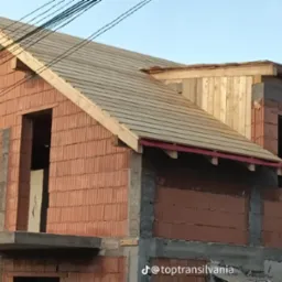 Budowa domu z czerwonej cegły: częściowo pokryty dach z drewnianą konstrukcją, widoczne ściany i otwory okienne. Na dole widoczny znak wodny.