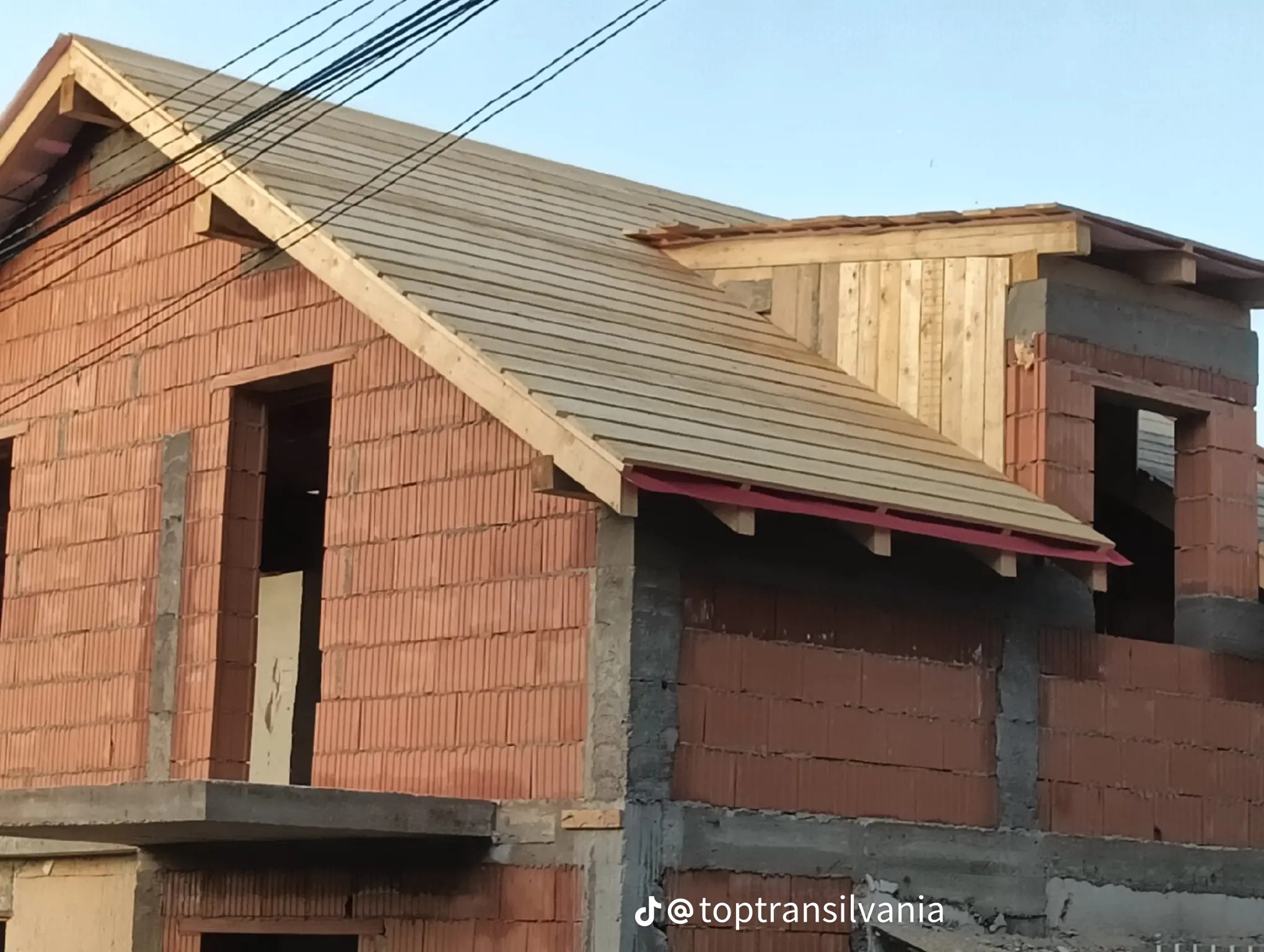 Budowa domu z czerwonej cegły: częściowo pokryty dach z drewnianą konstrukcją, widoczne ściany i otwory okienne. Na dole widoczny znak wodny.