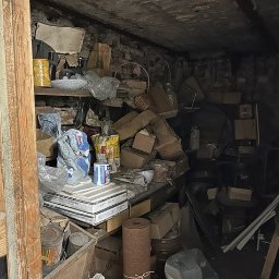 Tobiasz Czaplicki - Zagracone pomieszczenie z ceglanymi ścianami i półkami, wypełnione różnymi przedmiotami, kartonami i narzędziami. Widać stare puszki i zwitki materiału.