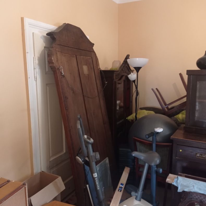 Zagracone pomieszczenie z meblami, sprzętem sportowym i kartonami przed opróżnianiem. Stara szafa oparta o ścianę, rower stacjonarny i piłka gimnastyczna.