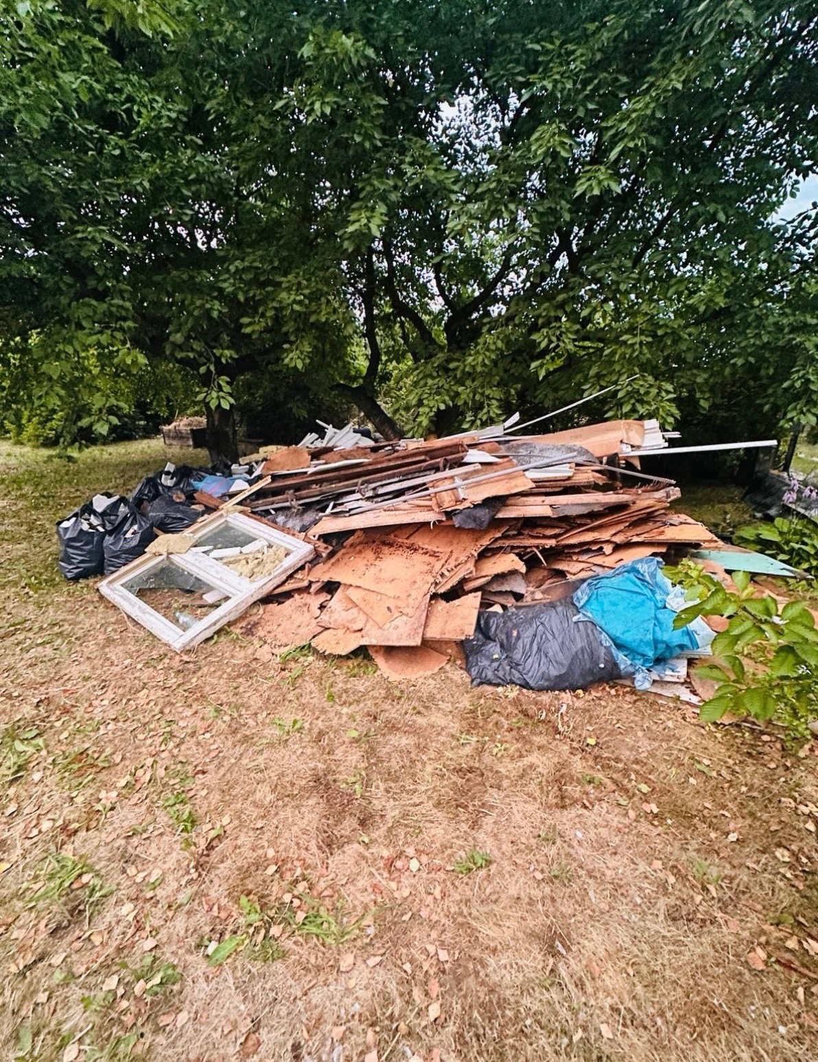 Sterta odpadów budowlanych i worków ze śmieciami na trawie, pod drzewem. Widoczne fragmenty okien, płyty i gruz. Opróżnianie po remoncie na zewnątrz.