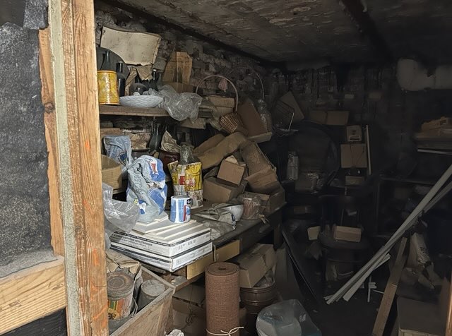 Zagracone pomieszczenie z ceglanymi ścianami i półkami, wypełnione różnymi przedmiotami, kartonami i narzędziami. Widać stare puszki i zwitki materiału.