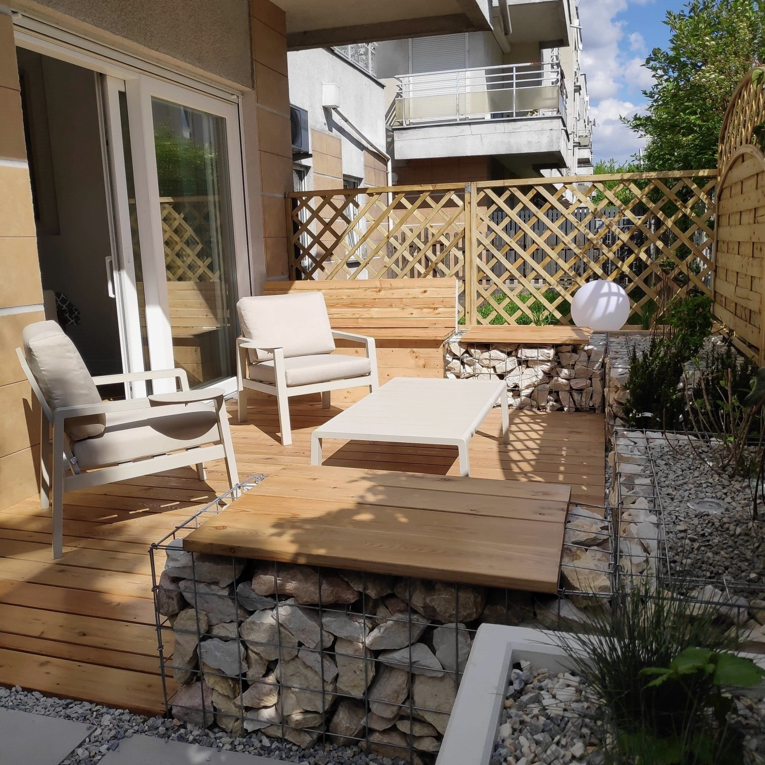 Minimalistyczny taras z drewnianą podłogą i gabionami. Dwa krzesła z poduszkami i stolik kawowy. Dekoracyjna lampa kula i ażurowe drewniane panele.