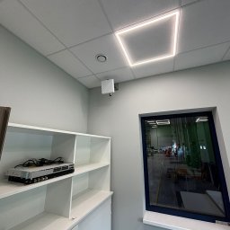 cenIT Andrzej Cendrowski - Białe wnętrze biurowe z półką na sprzęt sieciowy i oknem na halę produkcyjną. Widoczny switch sieciowy, punkt dostępowy WiFi i kwadratowa lampa sufitowa LED.