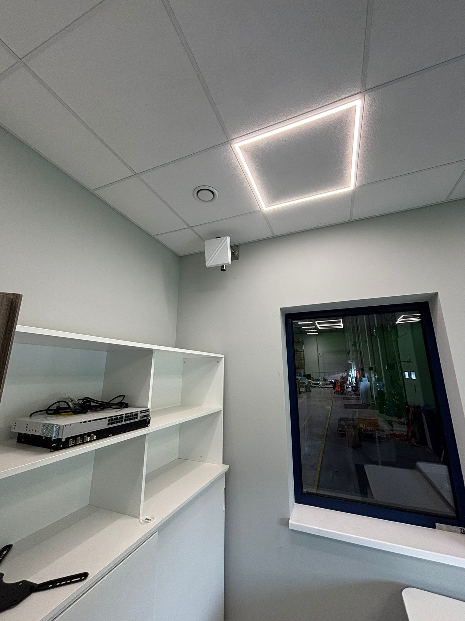 Białe wnętrze biurowe z półką na sprzęt sieciowy i oknem na halę produkcyjną. Widoczny switch sieciowy, punkt dostępowy WiFi i kwadratowa lampa sufitowa LED.
