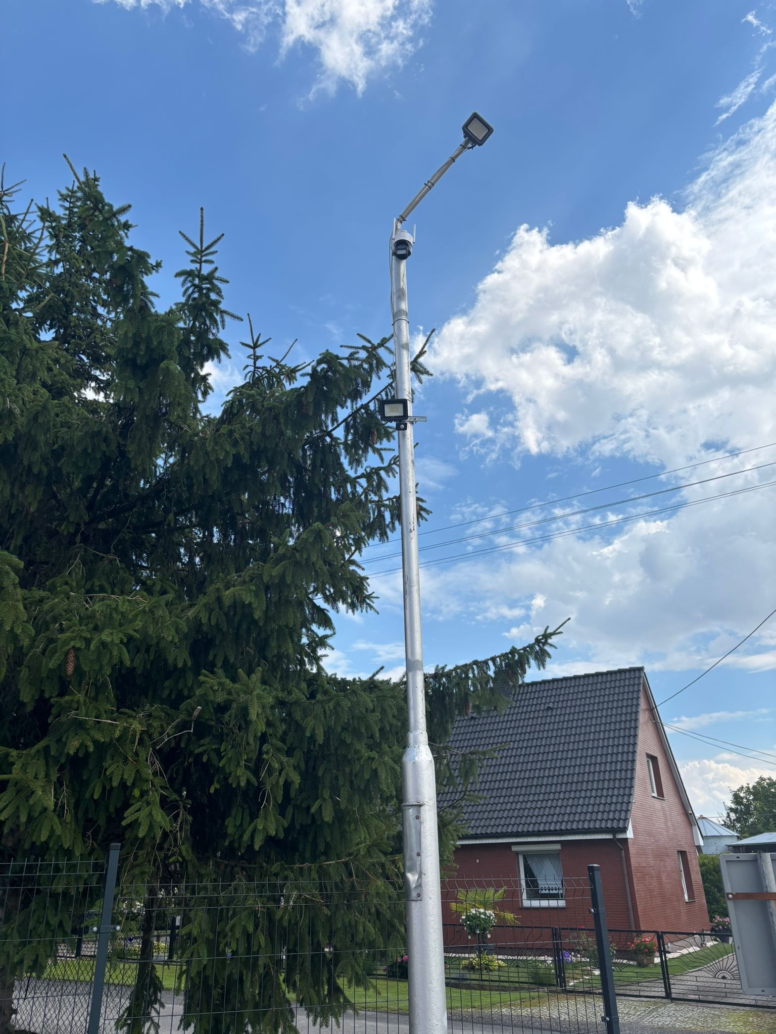 Srebrny słup oświetleniowy z kamerą monitoringu na tle błękitnego nieba i domu z ciemną dachówką, widoczny zza zielonego drzewa i metalowego ogrodzenia.
