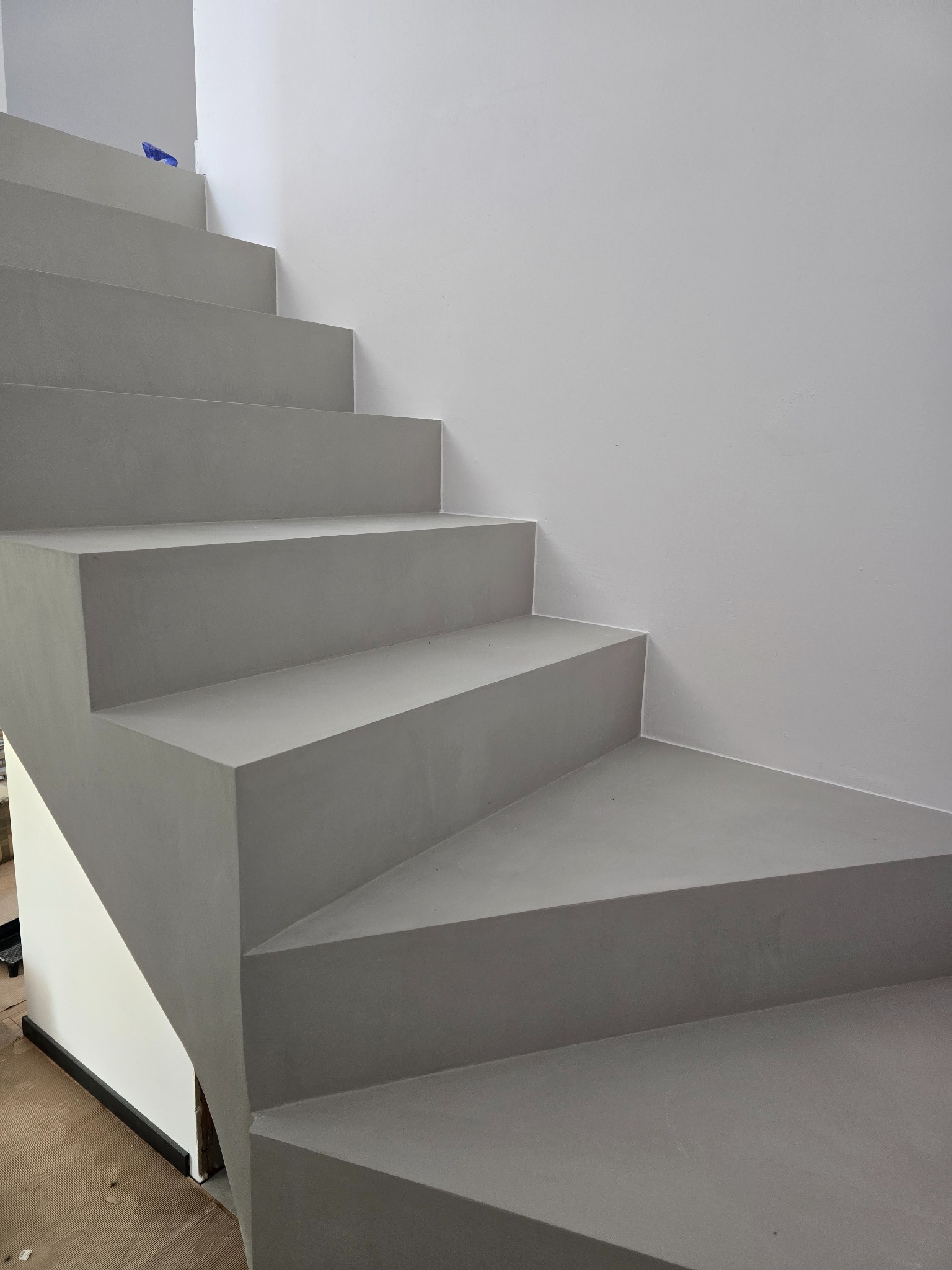 Schody pokryte szarym mikrocementem, gładka powierzchnia, minimalistyczny design, białe ściany, widoczne detale wykończeniowe u dołu schodów.