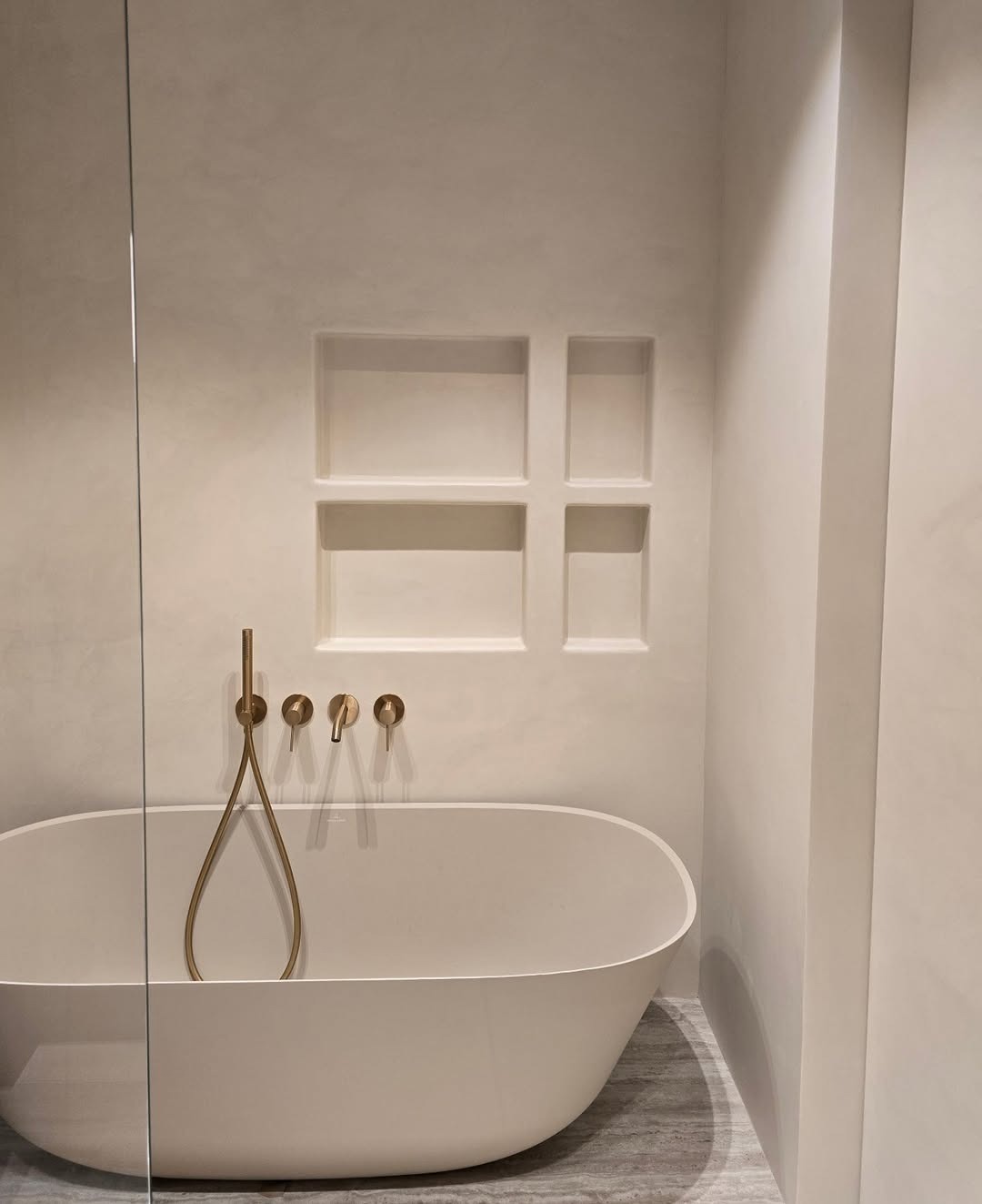 Minimalistyczna łazienka z owalną wanną, złotą armaturą i wnękami w ścianie wykończonej w jasnym, jednolitym kolorze. Widoczna szklana ścianka prysznicowa.