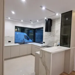 Jasna kuchnia na wymiar z marmurowym blatem i wyspą, czarny okap. Za oknem widoczny samochód dostawczy. Minimalistyczny design z oświetleniem punktowym.