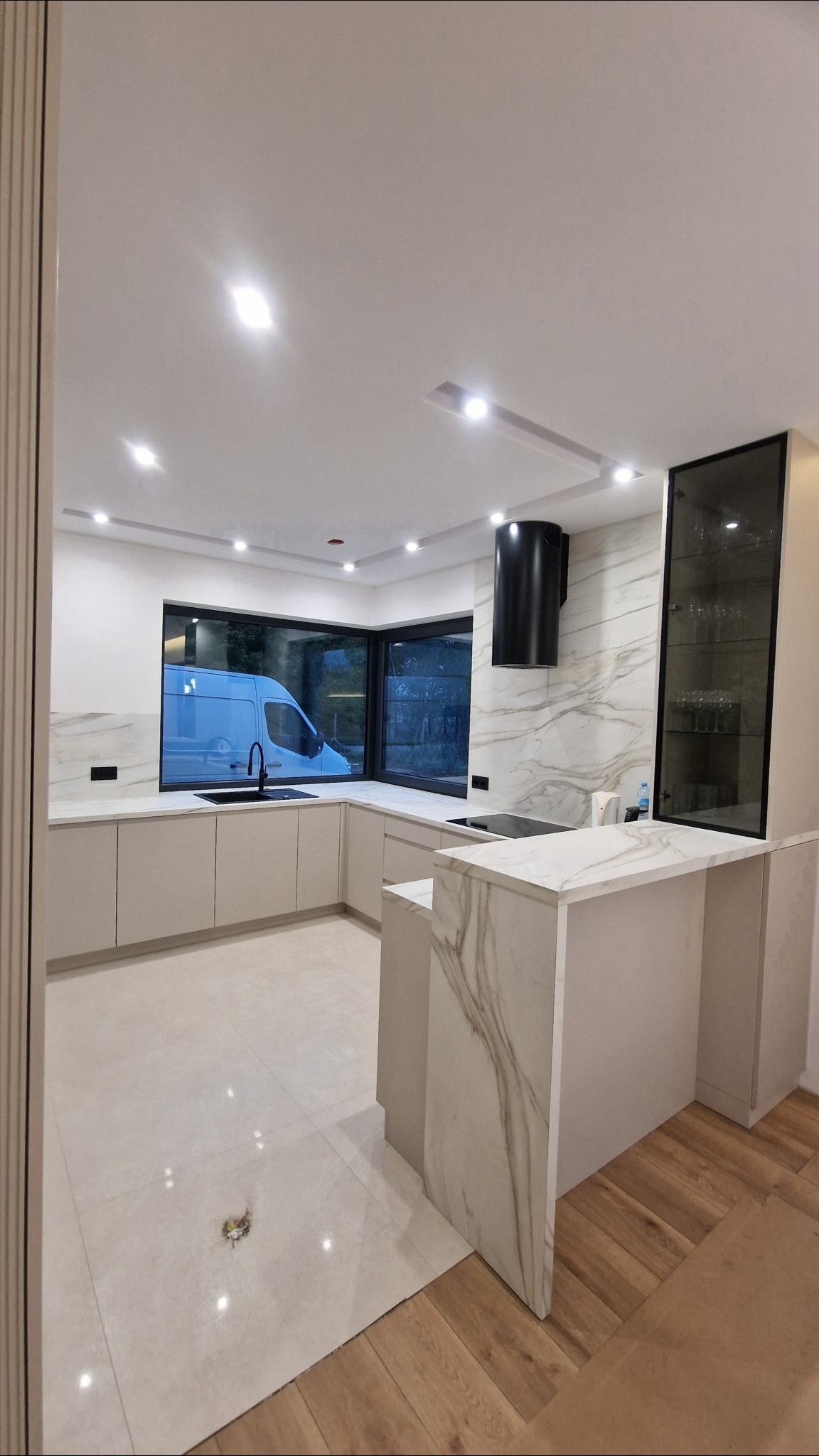 Jasna kuchnia na wymiar z marmurowym blatem i wyspą, czarny okap. Za oknem widoczny samochód dostawczy. Minimalistyczny design z oświetleniem punktowym.