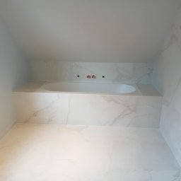 BLUTEX PAWEŁ SIWIŃSKI - Wykończona łazienka na poddaszu z wanną obudowaną płytkami imitującymi marmur. Widoczne przyłącza wodne i okno. Minimalistyczny design z przewagą bieli i szarości.