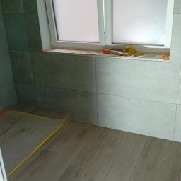 BLUTEX PAWEŁ SIWIŃSKI - Ułożone płytki podłogowe imitujące drewno w łazience z oknem. Widoczne narzędzia i materiały budowlane. Szare płytki ścienne tworzą spójną całość z podłogą.