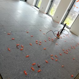 BLUTEX PAWEŁ SIWIŃSKI - Ułożone szare płytki podłogowe z pomarańczowymi klipsami poziomującymi w przestronnym, jasnym pomieszczeniu z oknami. Widoczny sprzęt glazurniczy.