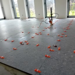 BLUTEX PAWEŁ SIWIŃSKI - Szare płytki podłogowe w trakcie układania, z pomarańczowymi klipsami poziomującymi. W tle widoczne okna i narzędzie glazurnicze.