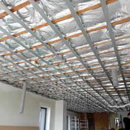 BLUTEX PAWEŁ SIWIŃSKI - Sufit podwieszany w trakcie montażu: metalowa konstrukcja, izolacja folią aluminiową, widoczne drewniane elementy. Prace wykończeniowe w pomieszczeniu.