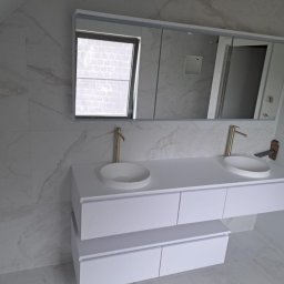BLUTEX PAWEŁ SIWIŃSKI - Wykończona łazienka w bieli: marmurowe płytki, podwójna umywalka z szafką i złotą armaturą, duże lustro z oknem. Nowoczesny design i dbałość o detale.