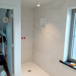 BLUTEX PAWEŁ SIWIŃSKI - Nowoczesna kabina prysznicowa z białymi płytkami imitującymi marmur i odpływem liniowym. Widoczne okno i otwór na głośnik w ścianie. Minimalistyczny design.