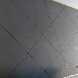 BLUTEX PAWEŁ SIWIŃSKI - Szara ściana z geometrycznym wzorem w romby, uzyskana techniką dekoracyjną. Widoczne delikatne nierówności faktury i linie podziału. Fragment drabiny w prawym dolnym rogu.