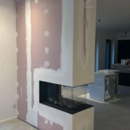 Wnętrze w trakcie prac wykończeniowych: obudowa kominka z płyt karton-gips, widoczne szpachlowanie i fragmenty niezabezpieczonej podłogi.