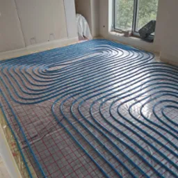 Niebieskie rury ogrzewania podłogowego ułożone na folii z czerwoną kratką w jasnym pomieszczeniu z oknem. Widoczne płyty kartonowo-gipsowe na ścianie.